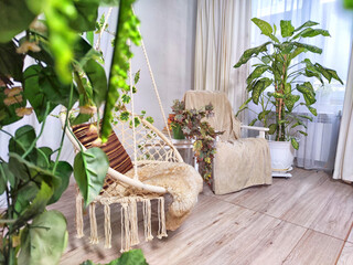 A modern cozy beautiful room with a braided rope macrame swing, chair, green plants, small table...