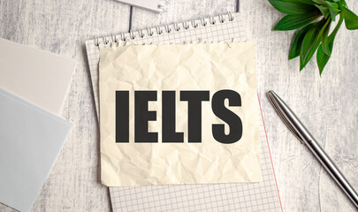 Crumpled paper with IELTS text on clipboard. Business concept with pen and green plant on wooden background. - obrazy, fototapety, plakaty