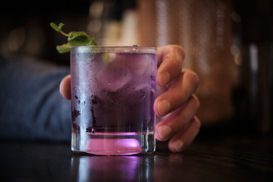 A close-up image capturing the allure of a refreshing purple cocktail adorned with a sprig of green mint, held by a person in a cozy, dimly lit setting