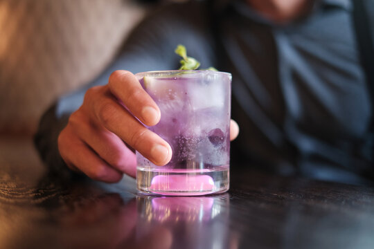 A close-up image capturing the allure of a refreshing purple cocktail adorned with a sprig of green mint, held by a person in a cozy, dimly lit setting
