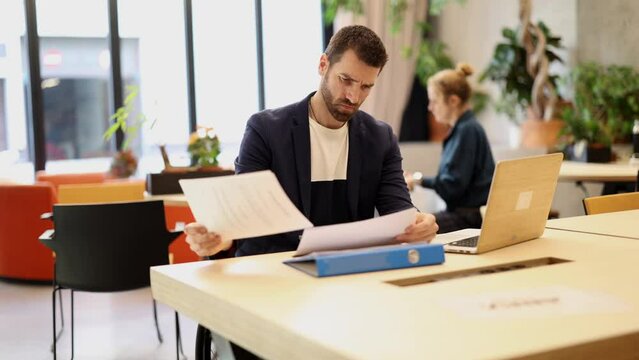 Focused Male Professional Reviewing Documents at Desk - A man intensely focused on paperwork with a laptop in a sunlit office space.