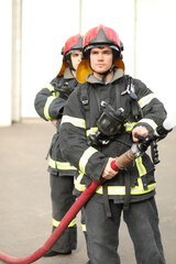 Obraz premium Portrait of two heroic fireman holds and adjust nozzle and fire hose spraying high pressure water, one is behind another