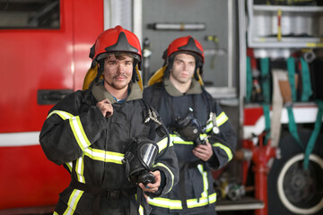 Two firefighters in protective suits and red fire helmet to protect head standing against fire engine
