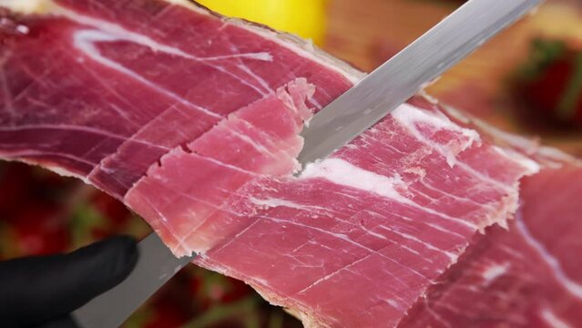 Red dry-cured prosciutto with some fat lines being sliced with sharp knife, traditional mediterranean delicacy for antipasto, process of slicing cured ham by professional chef
