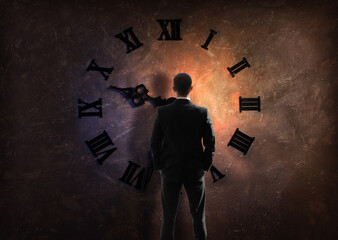 young businessman in black suit standing by wall with big clock with Roman dial, view from back