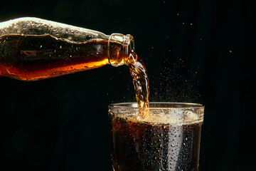 Soda, coke, dark beer pouring from bottle into glass against black background. Concept of alcohol...