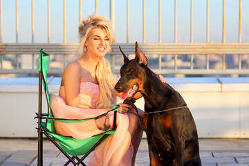Happy beautiful blonde in pink dress sitting on folding chair near the Doberman on the roof
