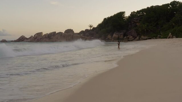 A woman standing on the beach while waves hit her legs. Beautiful rock formation in Seychelles, Africa. Huge waves crashing against the shore; boulders, warm sunset on an island; travelling alone