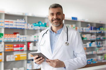 Middle aged male pharmacist druggist or doctor in white medical coat holding tablet with...