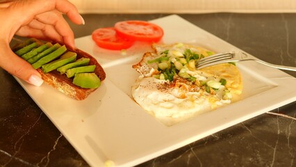 Woman places avocado sandwich on plate alongside omelet breakfast. Experience the richness of avocado in this wholesome meal. Witness avocado complementing the flavors of the omelet