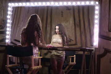Actress in shiny short dress looks in large makeup room mirror with lights around the perimeter
