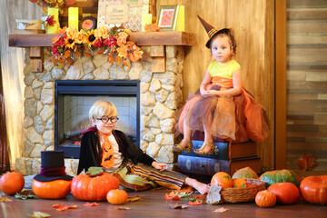 Little boy in black glasses with pumpkins near fireplace and girl in lush skirt sitting on pile of big books