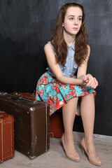 Portrait of young girl in floral skirt sitting on old suitcases in front of black wall