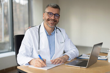 Happy middle aged man doctor in white medical coat, working at his workplace at modern clinic, writing prescription and using laptop computer, smiling at camera - obrazy, fototapety, plakaty