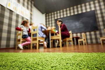 Children and nursery teacher sit at desks in classroom at child club, low angle view, focus on floor covering, shallow dof.