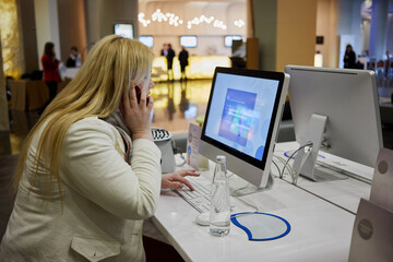 Blond woman talks on phone sitiing at desc with monitor. - obrazy, fototapety, plakaty