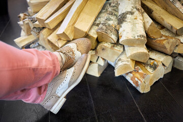 Female legs in pants and shoes near pile of birch firewood in room.