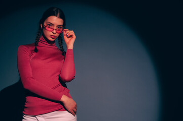 Fashionable confident woman wearing trendy pink sunglasses, turtleneck, posing on blue background. Studio fashion portrait. Copy, empty, blank space for text