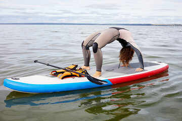 Woman in surfer suit stands in crab position on inflatable SUP board on water.