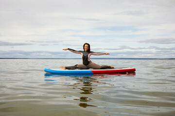 Young woman in surfer suit does split on inflatable SUP board on water.