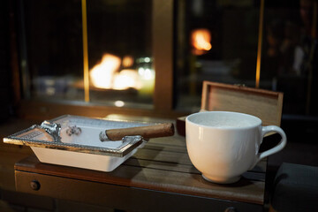 White cup of coffee, burning cigar on ashtray on wooden cigar table on veranda in evening.