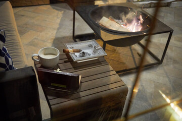 Small wooden cigar table with humidor, ashtray with burning cigar, cup of coffee near sofa and...