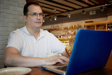 Fototapeta premium Man sits at table with laptop, cup of coffee and croissant in hand in cafe.