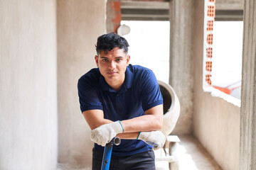 Portrait of confident young male mason leaning on shovel in corridor at incomplete construction site