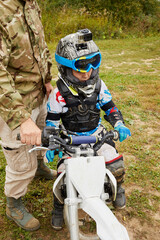 Little boy equipped and dressed for motorbike racing sits on motorcycle, man holds handle bar.