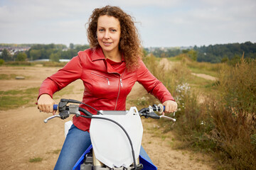 Woman in red leather jacket sits on motorbike outdoor on dusty track road.