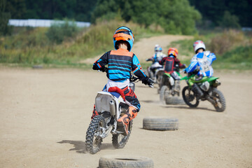 Four boys ride motorbikes between tyres lying on dusty ground.