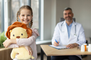 Portrait of happy girl patient holding toy and smiling at camera, posing in clinic office, male pediatrician in white coat on background. Professional medical services