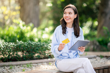 Radiant young woman sitting on a park bench holds a golden credit card and tablet