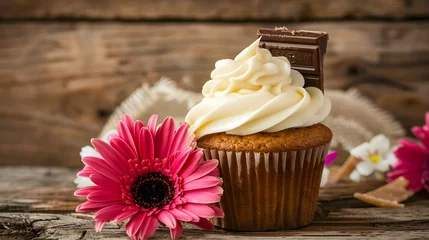 Tuinposter Buttercream topped cupcake Tasty cake with frosting and a miniature chocolate bar Sweet treat with a topping Placed on a rustic wooden background Decorated with gerbera flowers © PSCL RDL