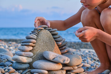 The child's hands lay pebbles, stones in a tower on the beach in summer, close-up.
