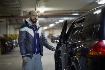 Bearded man stands opening door of modern black car at underground parking.
