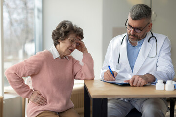 Obraz premium Sad senior woman sitting on examination and touching her stomach, suffering from pain and feeling unwell, man doctor writing prescriptions at appointment in clinic interior