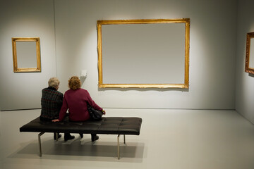 Two women sit on couch in room with frames on walls.