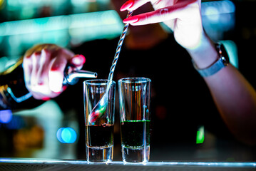 bartender pouring a dark liquid into two shot glasses at a bar, illuminated by colorful lights. The focus is on the pouring action, with the background blurred to emphasize this moment.