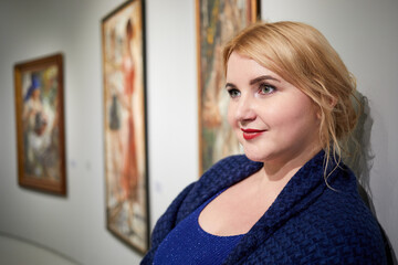 Smiling blond woman in blue dress stands against paintings on wall.