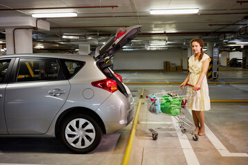 Young woman with shopping cart at underground parking near car with open trunk.