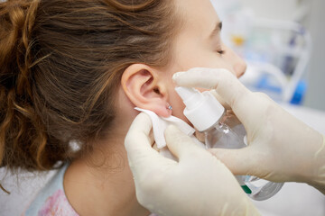 Doctor sterilize piercing place on ear of teenade girl.