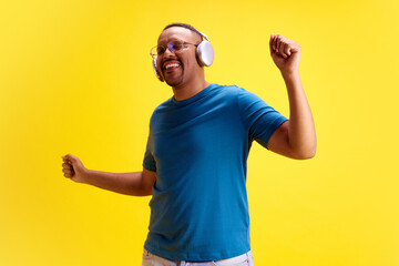 Cheerful, positive, relaxed African-American man in casual clothes listening to music in headphones and dancing against yellow studio background. Concept of human emotions, casual fashion, lifestyle