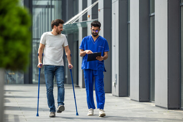 Young bearded man with crutches walking and talking to a male nurse