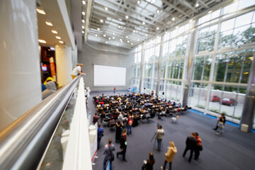 People listen to speaker in spacious auditorium, blurred image.