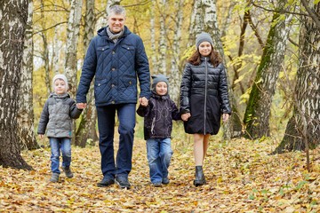 Father, daughter and two little sons walk down pathway in autumn park holding hands.