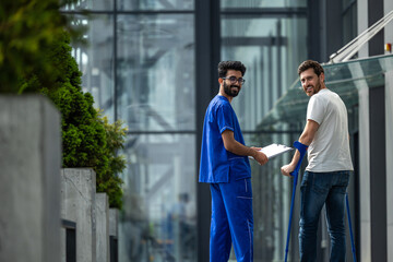 Man with crutches and a male nurse in a blue uniform walking in a hospital yard