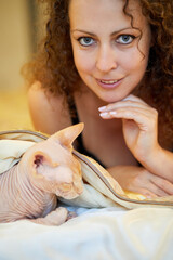 Smiling woman with curly hair and sphinx cat on bed.