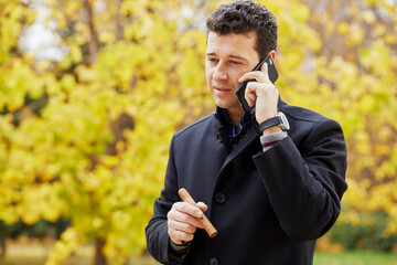 Half-length portrait of man with cigar talking cell phone in autumn park.