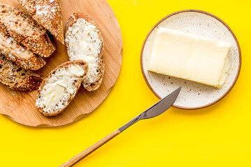 Wholegrain bread on cutting board with butter on plate, slice of bread with butter on yellow background top view